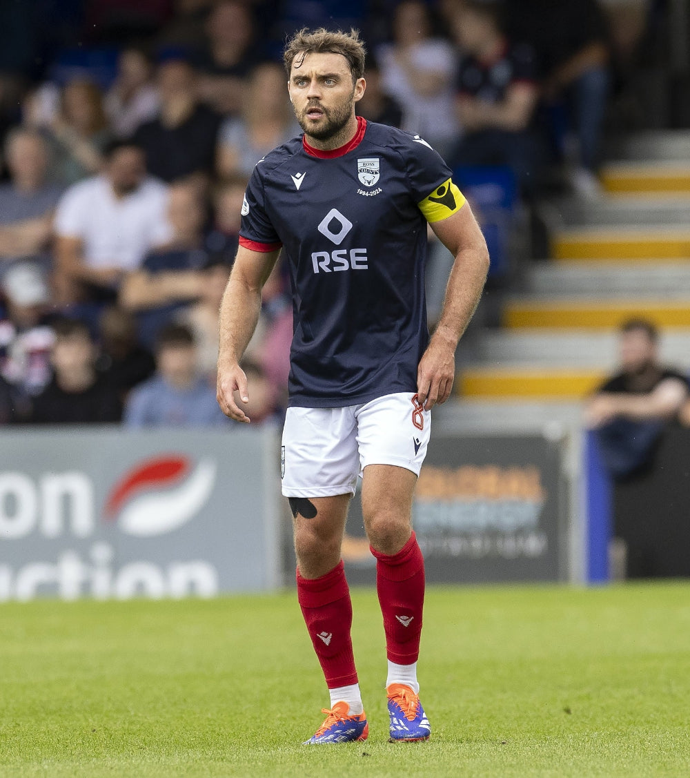 Soccer player in a dark blue jersey with 'RSE' on it, standing on a grass field with blurred spectators in the background.